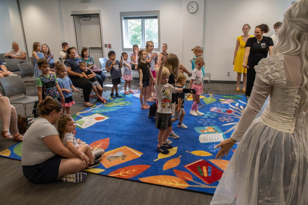 A group of children participating in a story-time event, with some standing and actively engaging on a colorful carpet while adults watch from the background.