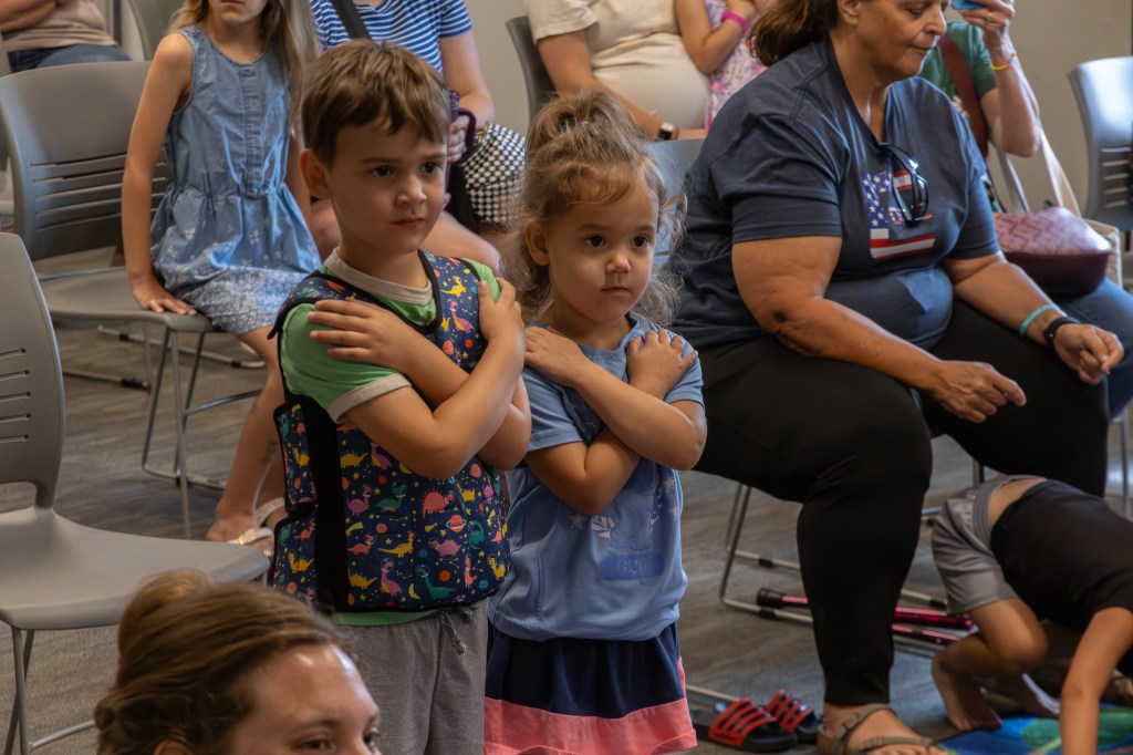 Two children, a boy and a girl, standing with their arms crossed in a seated audience during an event. Other attendees, including adults, are visible in the background.