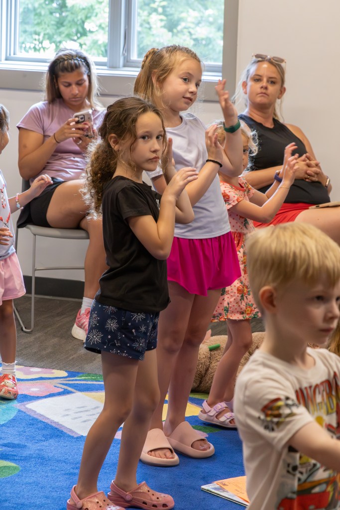 Two young girls participating in an activity, clapping their hands, while other children and adults observe in a bright, colorful room.