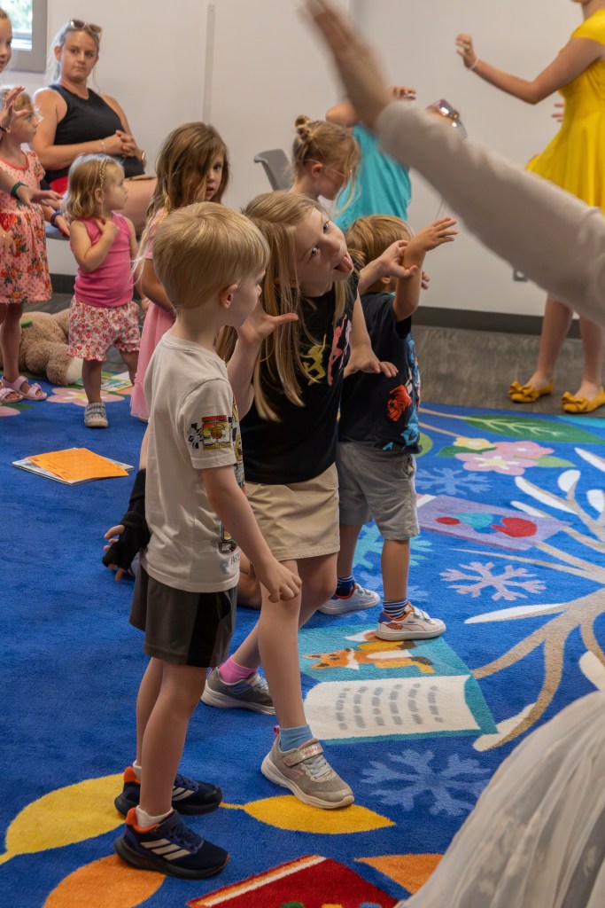 A group of young children participating in an interactive activity on a colorful rug, with attentive adults in the background.