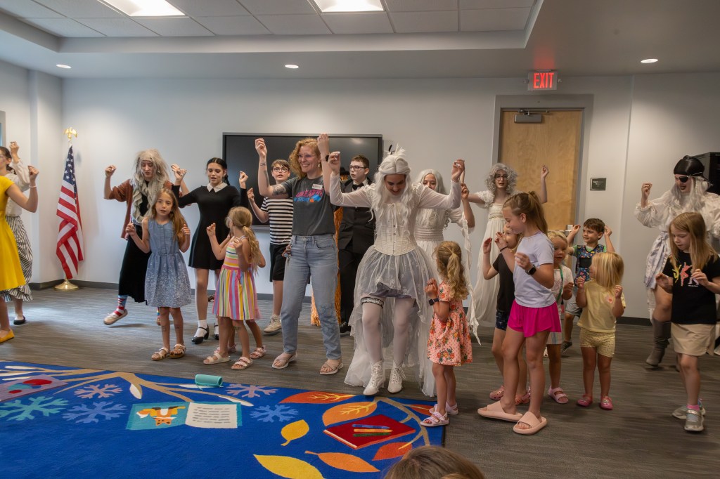 A group of children and adults dressed in costume are joyfully dancing together indoors. Some participants wear whimsical outfits, including a character in white and others in colorful attire. The setting includes an American flag in the background and a vibrant carpet design.