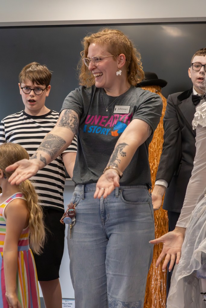 A smiling woman in a gray t-shirt with the text 'UNEARTH A STORY' is engaging with a group of children and adults during an interactive event. She is standing in the foreground, arms outstretched, while others look on enthusiastically.