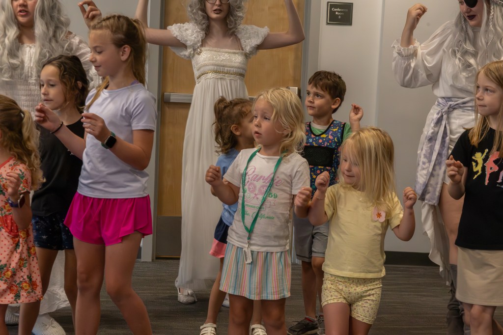 A group of children and adults participating in a playful activity, with some wearing costumes. The children are dancing and having fun in a conference room setting.
