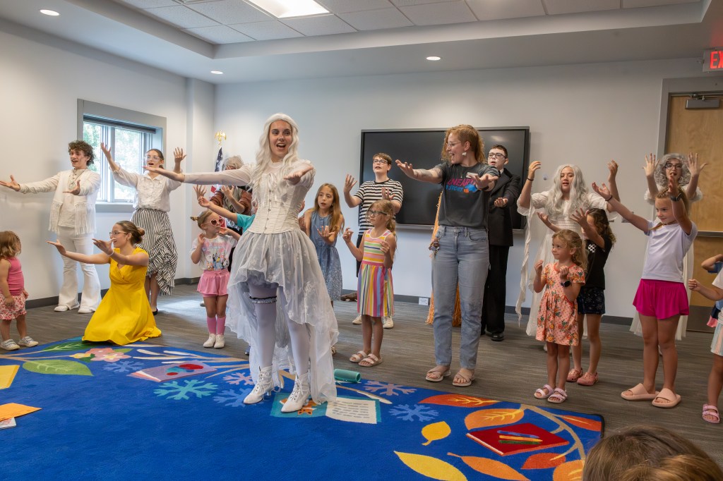 A group of children and performers in costumes participating in a theatrical performance inside a bright, cheerful room, with colorful carpeting and a screen in the background.