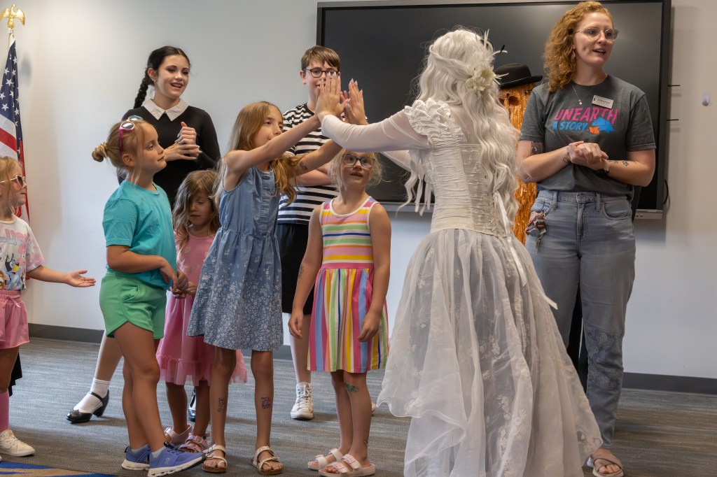 A group of children interacting with a character dressed in a white costume, while adults observe. The scene includes a U.S. flag in the background.