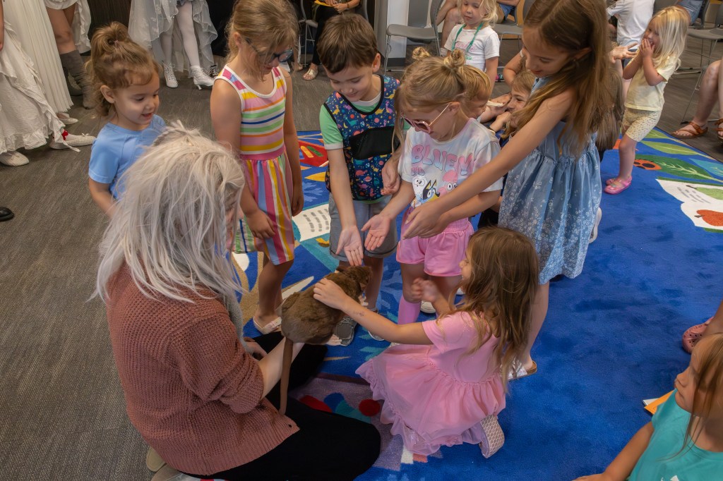 A group of children gathered around an adult, who is holding a small animal. The children are eagerly reaching out to pet the animal while sitting on a colorful rug.