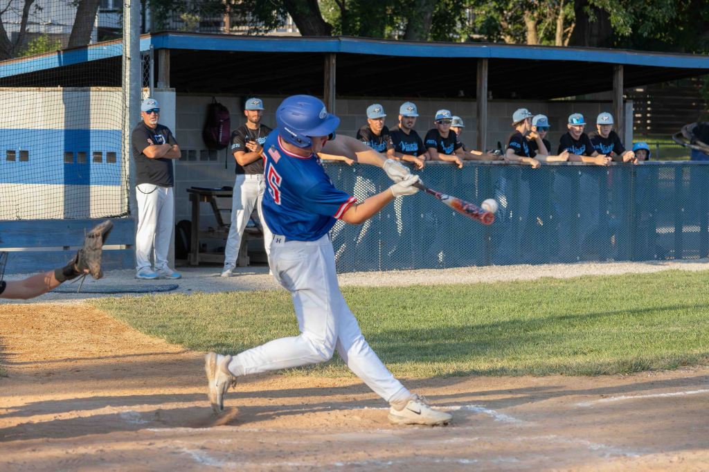 A young baseball player in blue attire swings a bat at a pitch, with spectators in the background watching from the dugout.