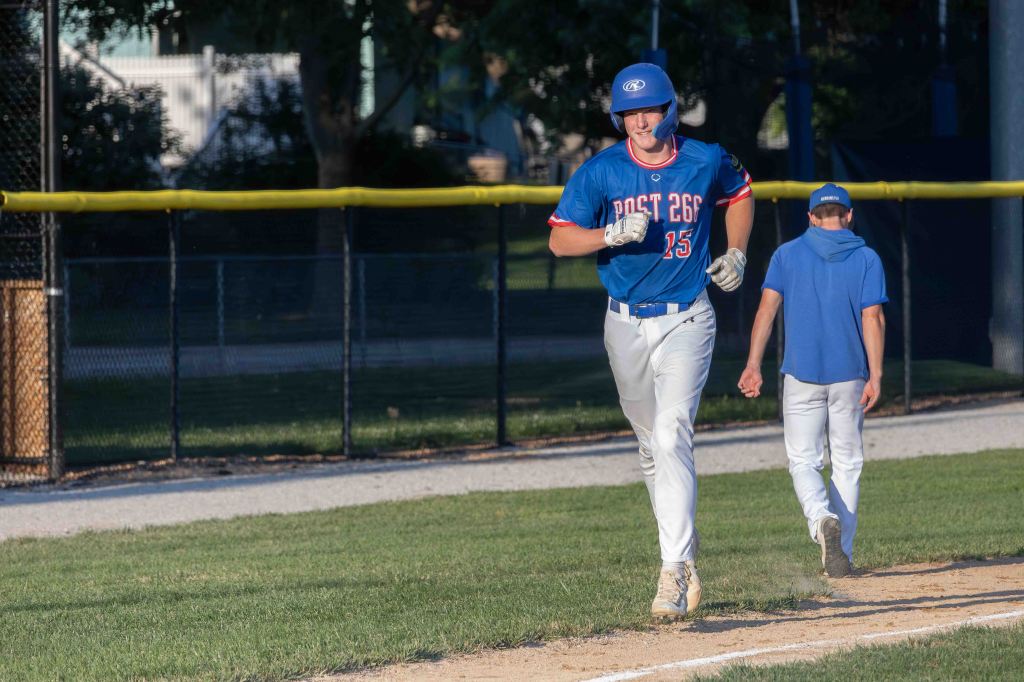 A baseball player in a blue jersey with 'Post 26' and the number 15 running along the base path, with another player in a blue shirt walking behind him.