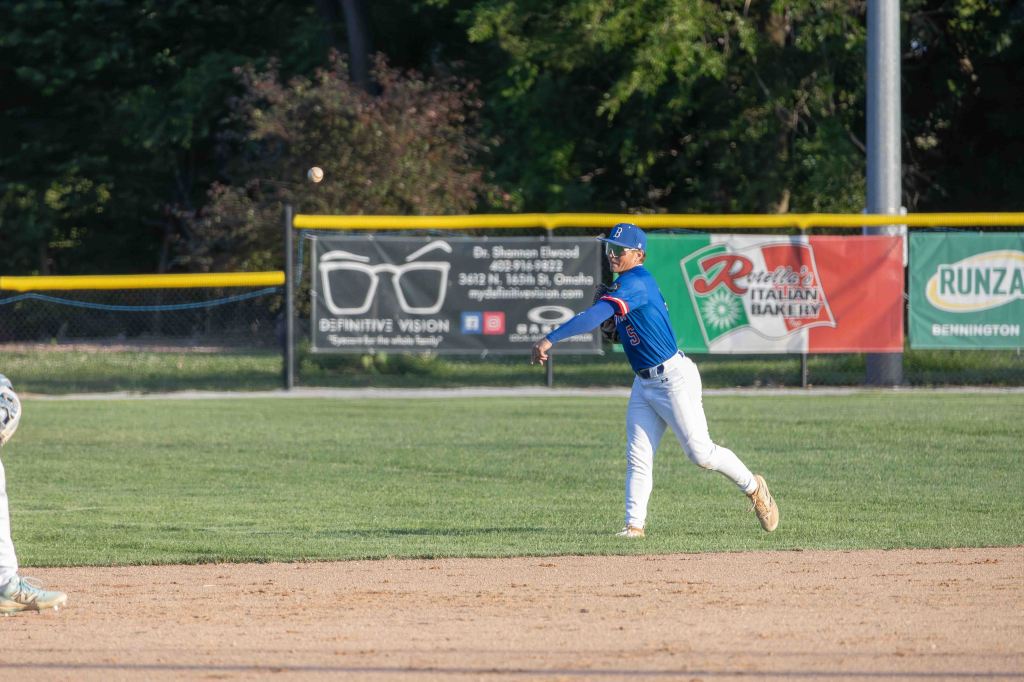 A young baseball player in a blue jersey and white pants is making a throw from the infield during a game, with baseball fields and advertisements in the background.
