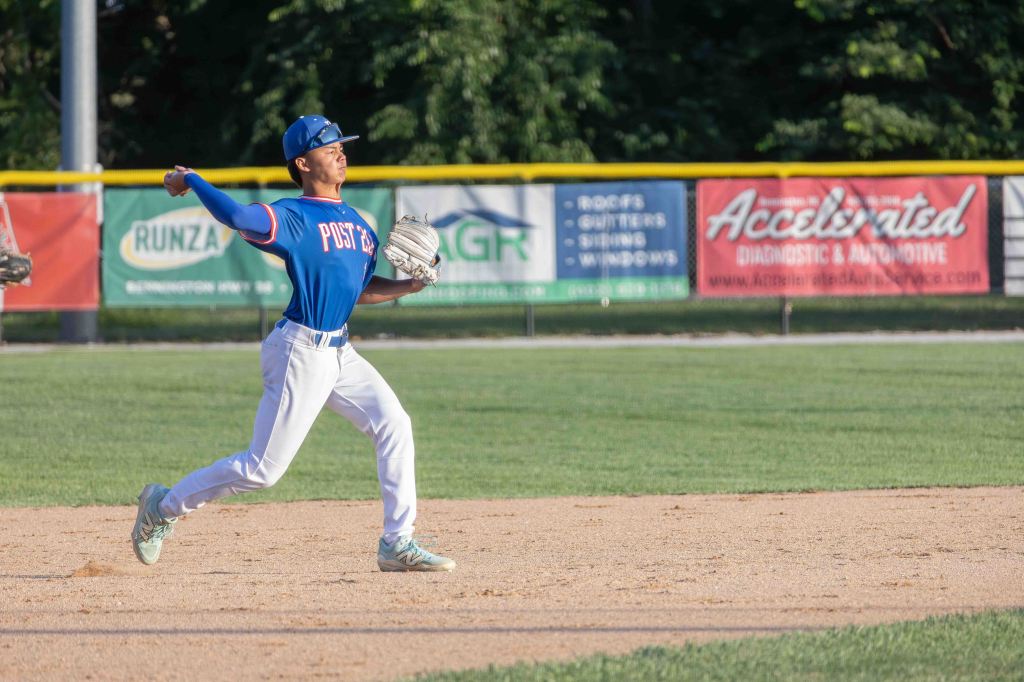 A young baseball player in a blue jersey with 'POST 22' printed on it, throwing a baseball on an infield during a game.