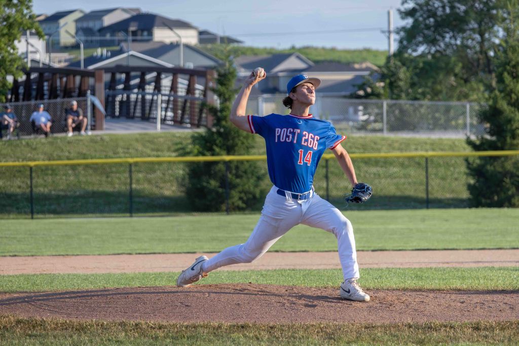 A young baseball pitcher in a blue jersey with the number 14 and 'POST 266' on it, in the midst of a throwing motion on a baseball field.