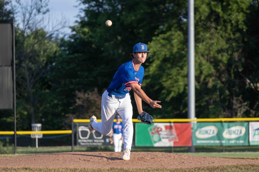 A young baseball pitcher in a blue jersey and cap throws a ball from the pitcher's mound during a game, with a blurred background of trees and a baseball field.