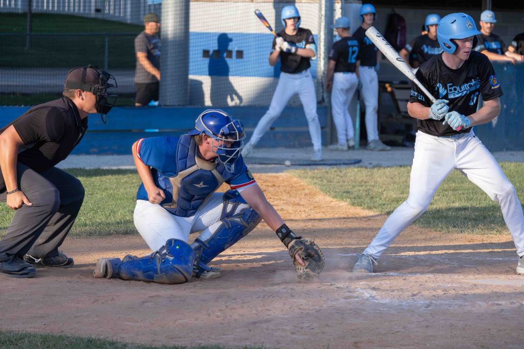 A baseball scene featuring a catcher in blue gear, an umpire in black, and a batter preparing to hit the ball. Players in the background are positioned on the field.