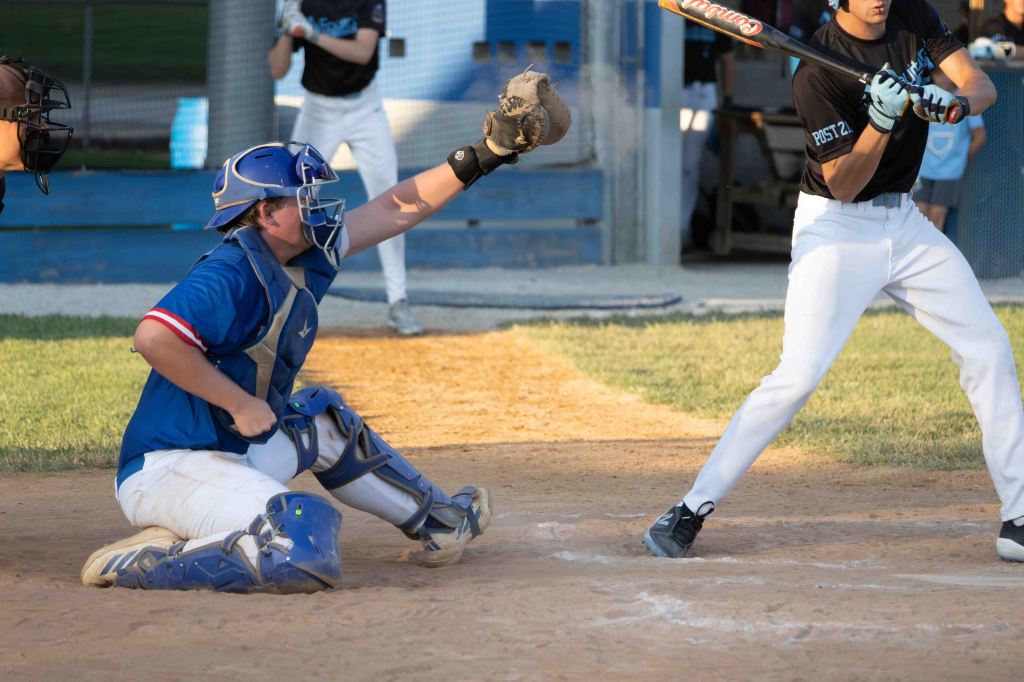A baseball catcher in a blue uniform stands ready to receive a pitch, while a batter in white prepares to swing. The scene is set on a dirt baseball field, with players in the background.