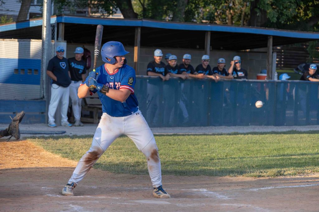 A young baseball player standing in a batting stance, preparing to hit a pitch, with a team of players in the background watching from the dugout.