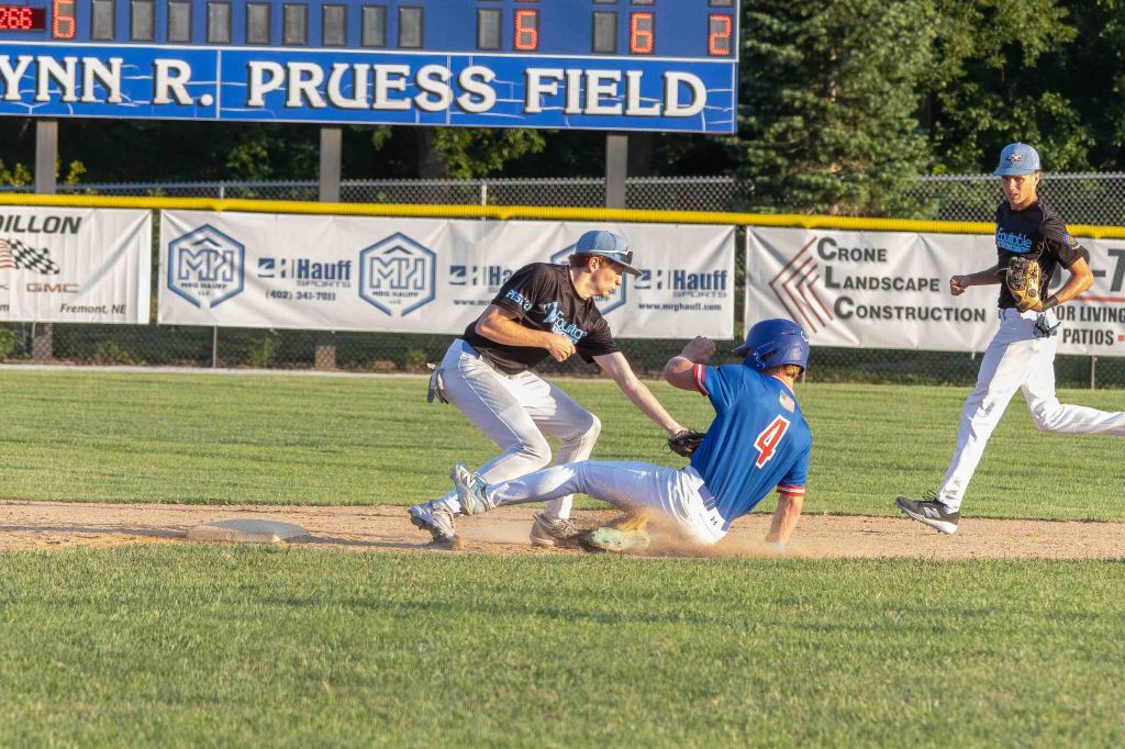 A baseball player sliding into base while being tagged by a defensive player, with two other players in the background and a scoreboard in the distance.