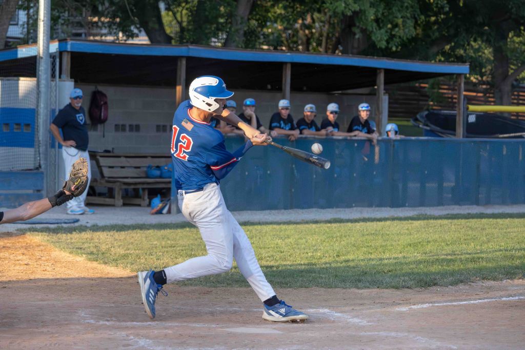A young baseball player in a blue jersey swings a bat at a ball during a game, while teammates watch from the dugout in the background.