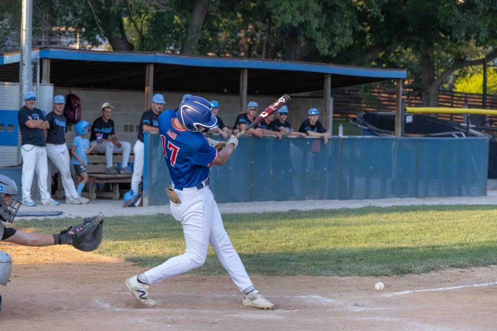 A youth baseball player swings a bat at home plate while a catcher crouches nearby. In the background, teammates watch from the dugout.