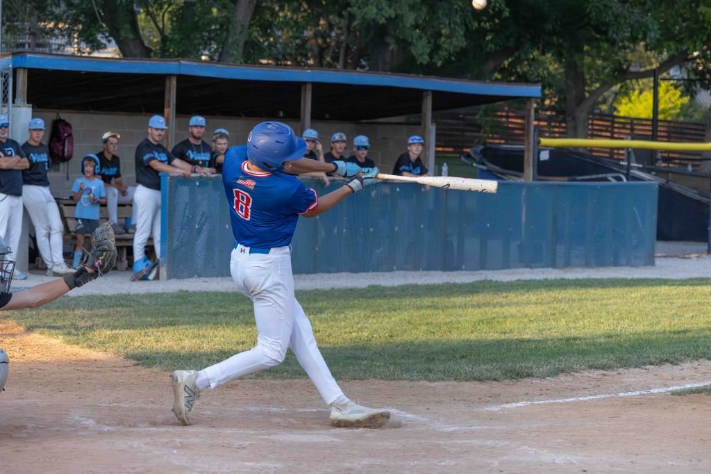 A young baseball player swings a bat during a game, with players in the dugout watching in the background.