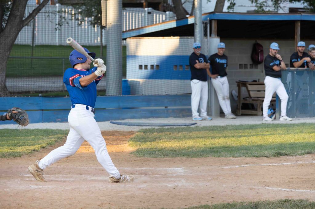 A baseball player in a blue jersey swings a bat at the baseball field, while teammates watch from the dugout in the background.