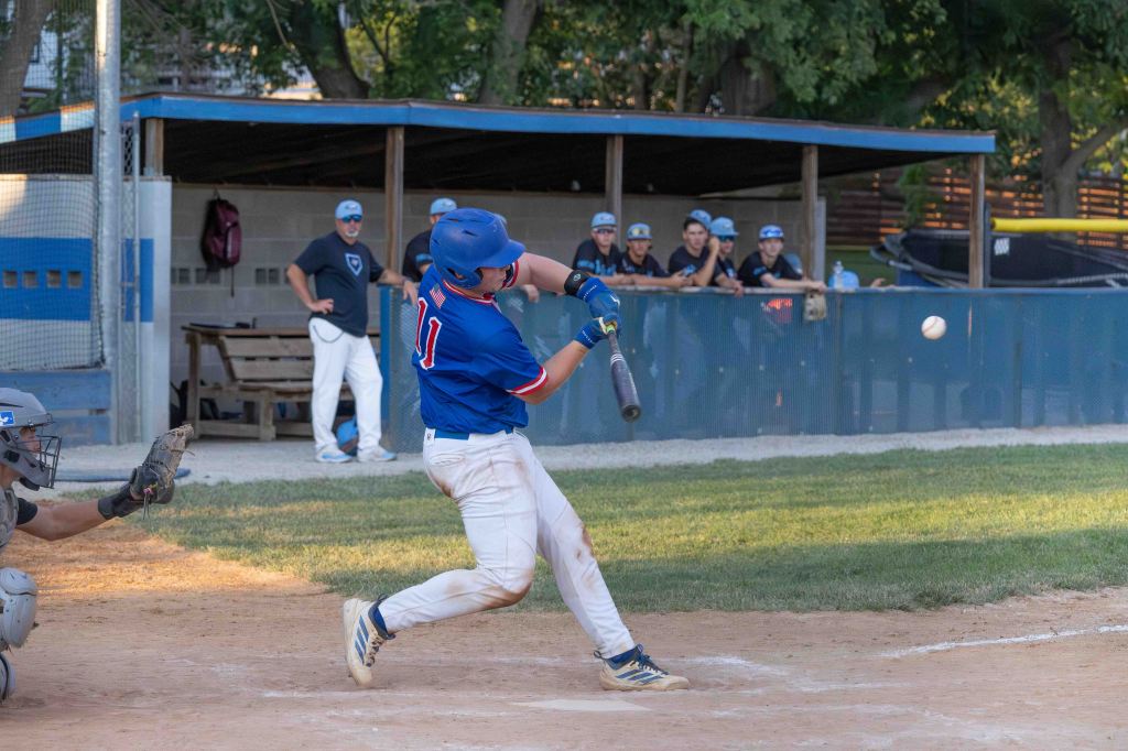 A baseball player in a blue jersey swings at a pitch while a catcher prepares to receive the ball. Teammates watch from the dugout in the background.