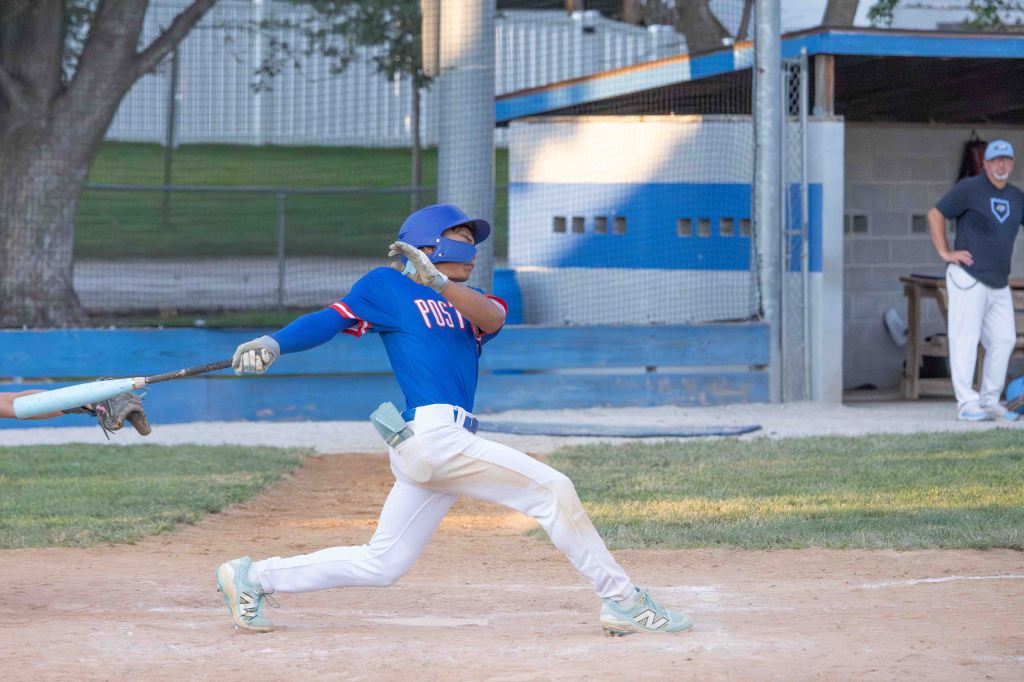 A young baseball player in a blue jersey swings a bat while batting in a game, with a coach visible in the background.
