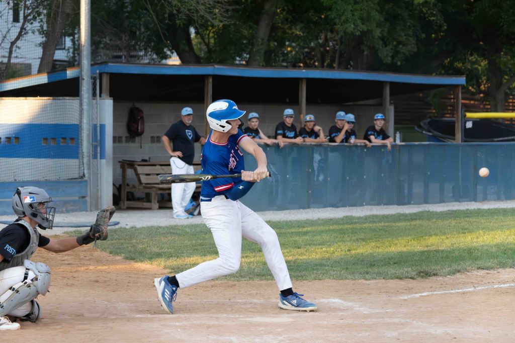 A young baseball player swings a bat during a game, while a catcher prepares to receive a pitch. In the background, teammates watch from the dugout.