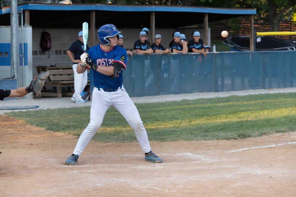 A baseball player in a blue jersey prepares to hit a pitched ball while another player crouches behind home plate. In the background, a group of teammates observes the play from the dugout.