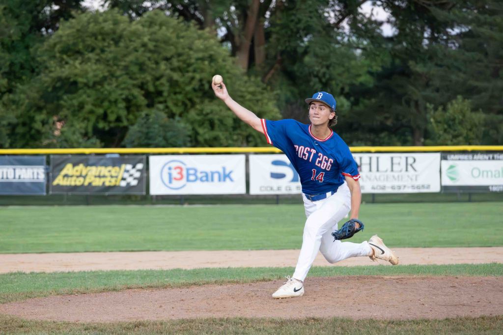 A young baseball player wearing a blue jersey and white pants is in a pitching motion, preparing to throw a baseball on a grassy field.