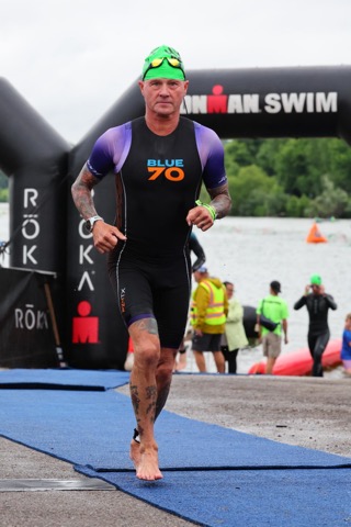 A male athlete wearing a wetsuit and goggles runs on a blue mat towards the finish line of an Ironman swim event, with spectators and kayakers in the background.