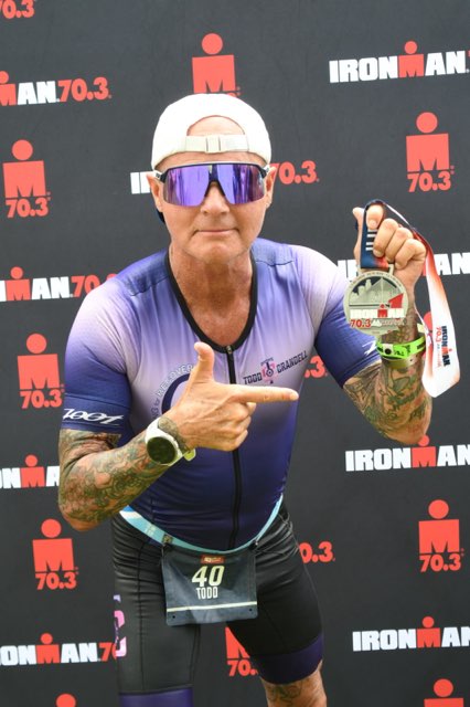 A middle-aged male athlete in a blue and purple racing suit is holding a medal and pointing at it, with a backdrop displaying the IRONMAN 70.3 logo.