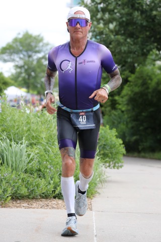 A male athlete in a purple and black triathlon suit runs along a path surrounded by greenery, wearing sunglasses and a race bib number 40 on his waist.