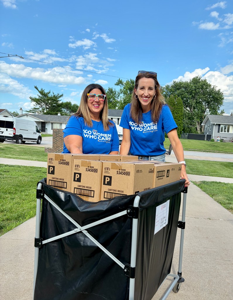 Two women smiling and standing next to a cart filled with cardboard boxes, wearing blue t-shirts that say '100 Women Who Care.'