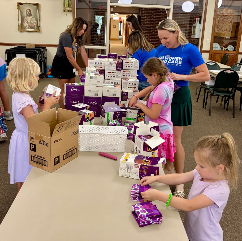 Young children and adults organizing packages on a table in a well-lit room, with boxes of products stacked in the background.