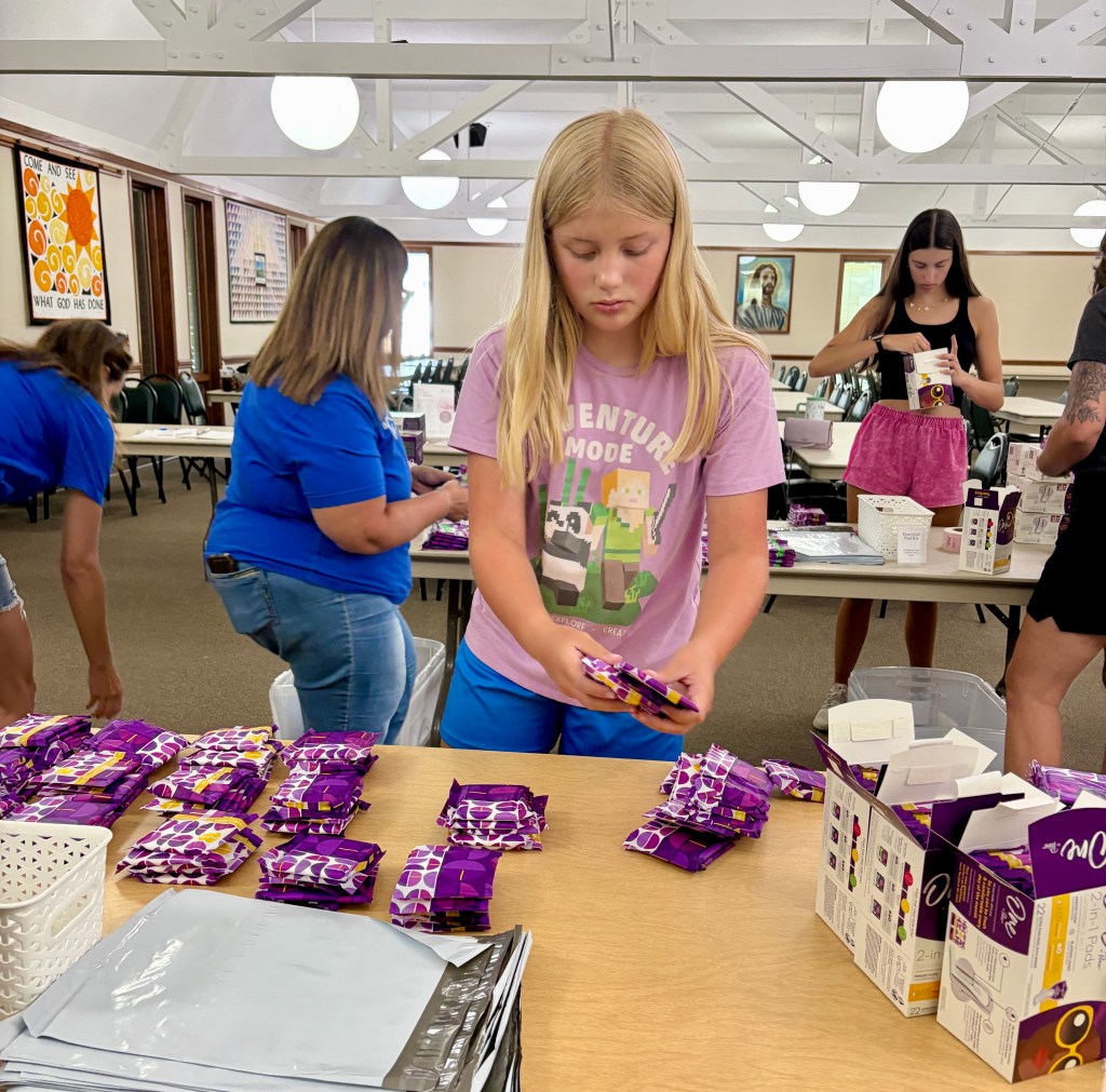A young girl with long blonde hair wearing a light pink t-shirt is organizing purple packages on a table in a community hall. In the background, two other people are also engaged in similar tasks.