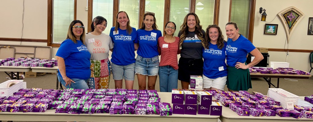 A group of seven women wearing blue shirts with the text '100 Women Who Care' posing together in front of tables filled with neatly arranged purple boxes and packages.