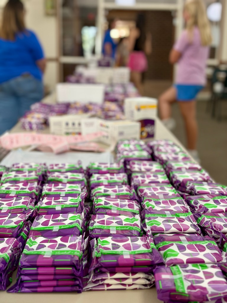 A table filled with stacked packages of purple and green products, likely personal hygiene items, with people in the background engaging in activities.