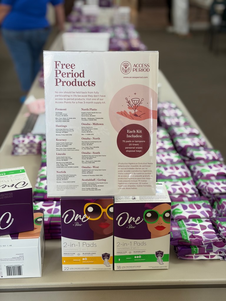 Display of free period products information, including a sign detailing access points and kits on a table, with menstrual pads and personal care items.