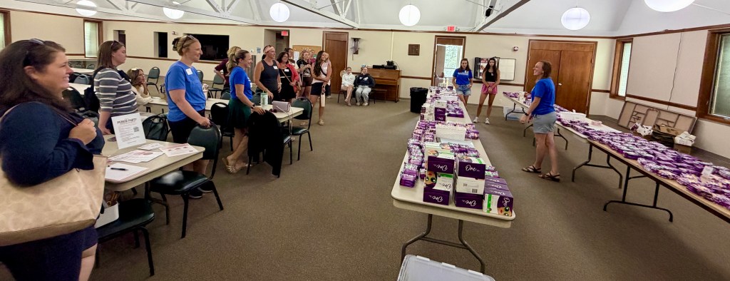 A gathering of participants in a community meeting room, some women wearing blue shirts are standing at the front with tables displaying products, while others are seated and engaged in conversation.