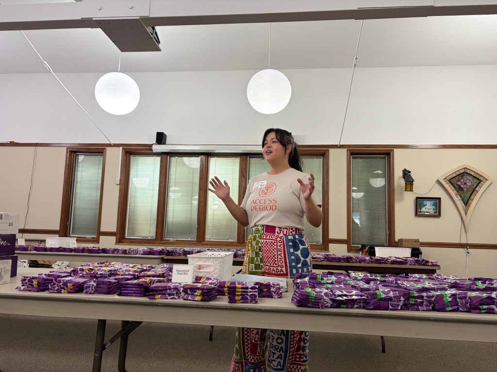 A woman wearing a shirt that says 'ACCESS PERIOD' stands in front of tables filled with purple packages while giving a presentation.