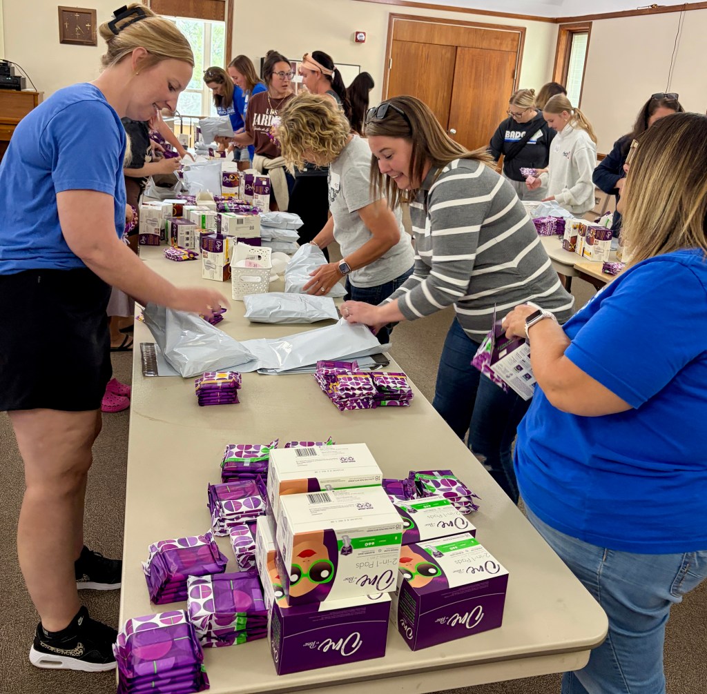 A group of women working together at tables to package products, including boxes of pads and sealed bags. The setting appears to be a community space with large windows and a friendly atmosphere.