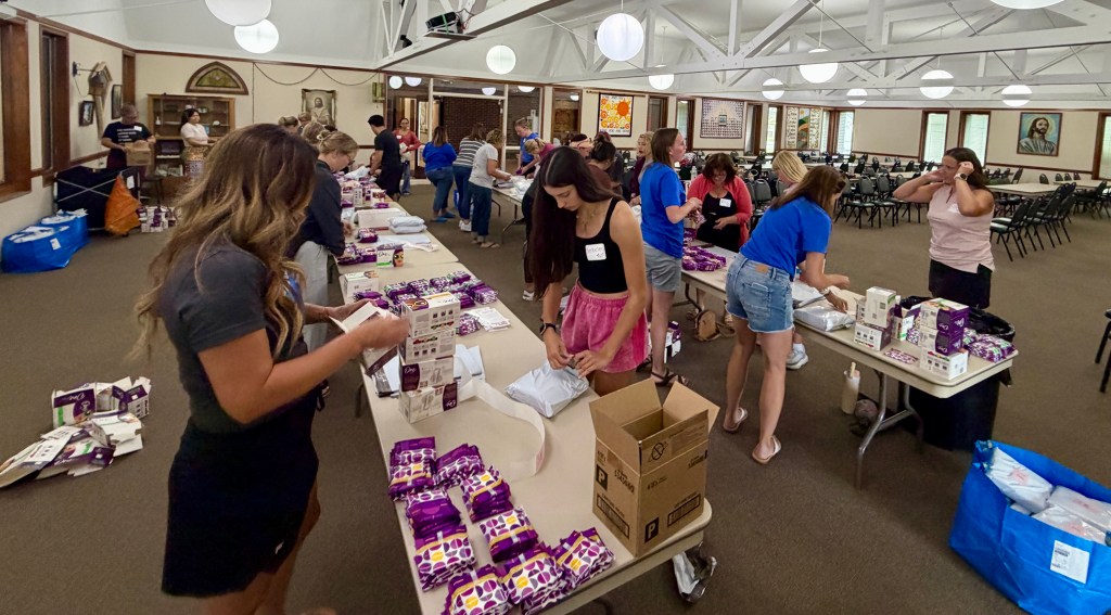 A group of volunteers packing items at tables in a spacious indoor hall. Some individuals are assembling boxes while others organize products in purple packaging.