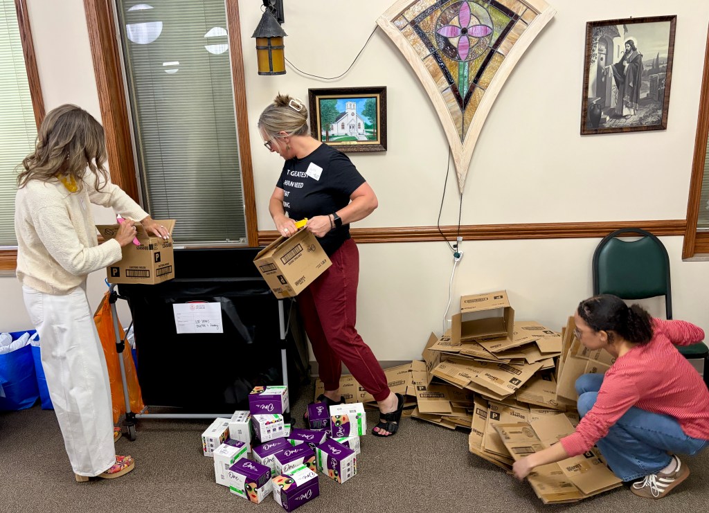 Three women sorting and recycling cardboard boxes in a community space, with one woman holding a box and another writing on it.