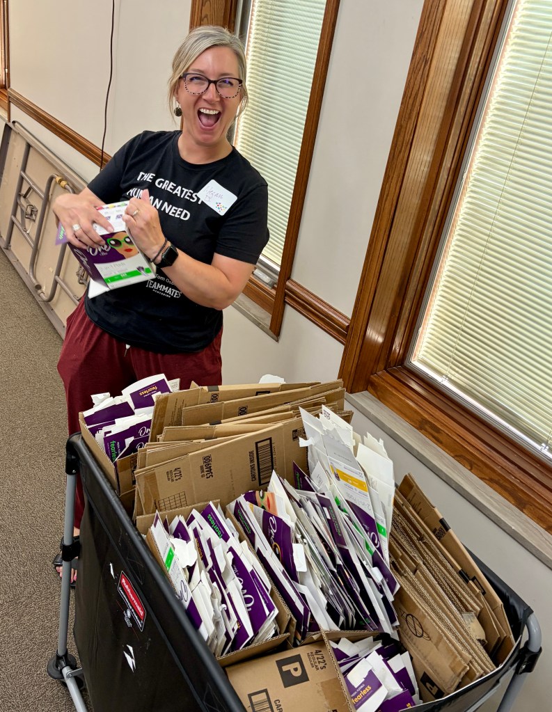A smiling woman wearing a black t-shirt poses next to a bin filled with various packages and cardboard boxes, showcasing an enthusiastic and playful demeanor.