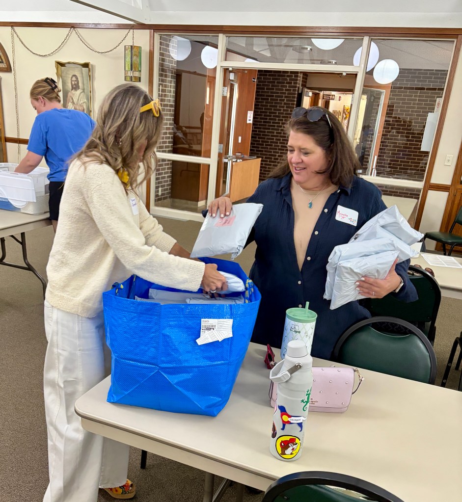 Two women packing items into a large blue tote bag during a gathering, with a table and other attendees in the background.