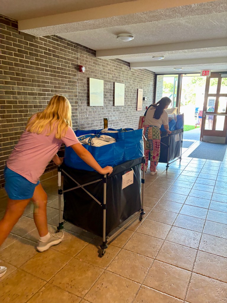 Two individuals pushing rolling carts filled with bags in a hallway with brick walls and a door leading outside.