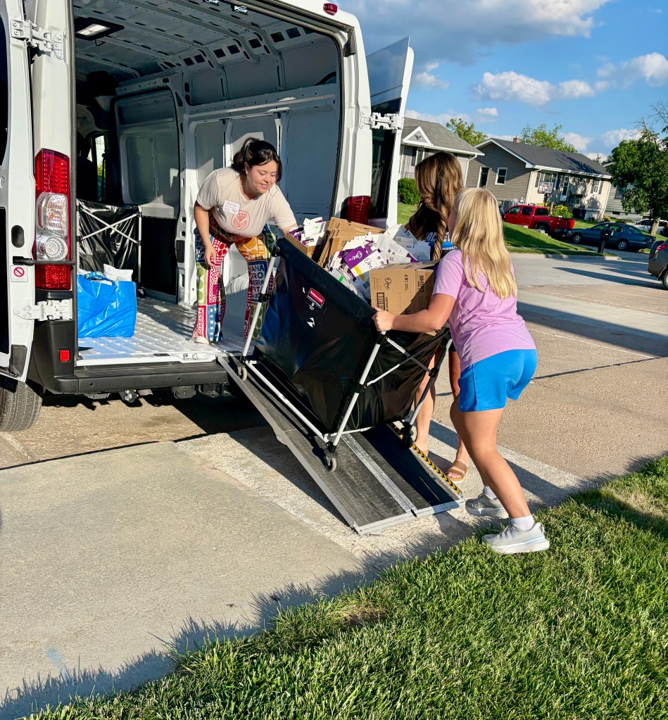 Two women and a girl unloading items from a van using a wheeled cart, with a sunny residential backdrop.