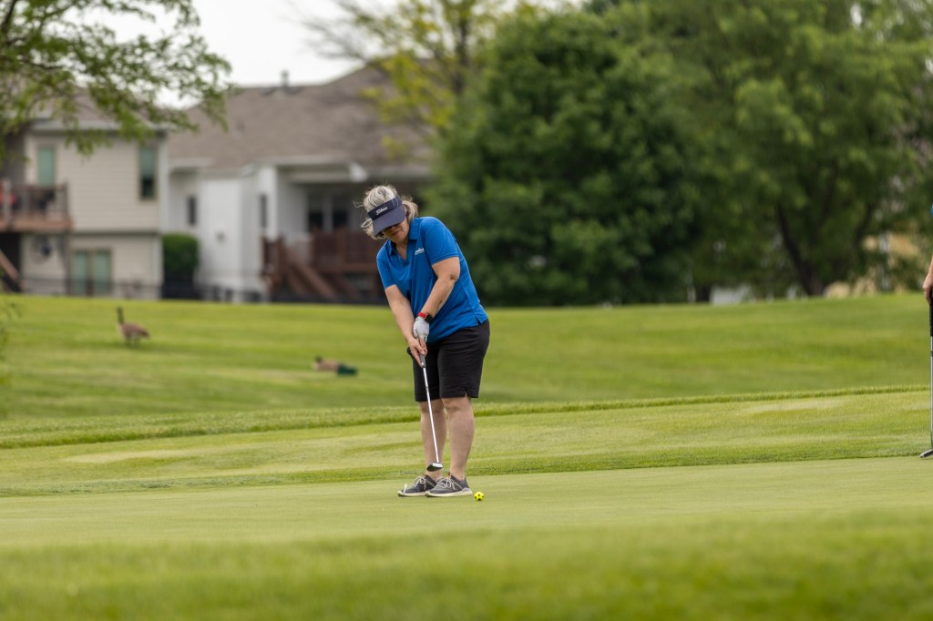 A golfer in a blue shirt and black shorts is putting on a green golf course, with residential houses and trees in the background.