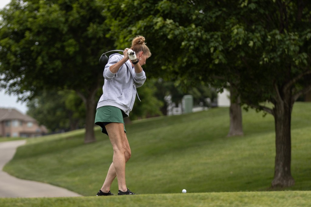 A woman dressed in a grey hoodie and green shorts prepares to hit a golf ball on a lush, green golf course.