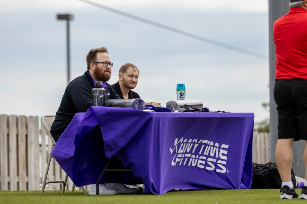 Two individuals seated at a table with a 'Anytime Fitness' banner, displaying promotional materials and a water bottle, outdoors on a grassy area.